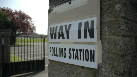 BBC a sign saying Way in polling station. The sign is raped to a stone wall and a railing and some parkland can be seen in the background