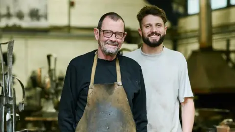 Brian Fell (L) has short cropped brown hair, brown glasses and wears a black sweat shirt and stained khaki apron while George Fell (R) has brown hair and beard and wears a grey T-shirt.
