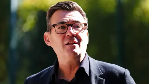 Andy Burnham wearing a dark suit and glasses.