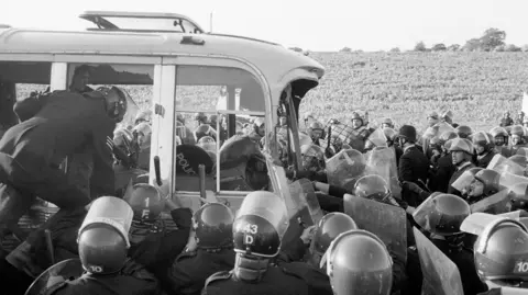 A black & white photo of police in riot gear surrounding an old bus