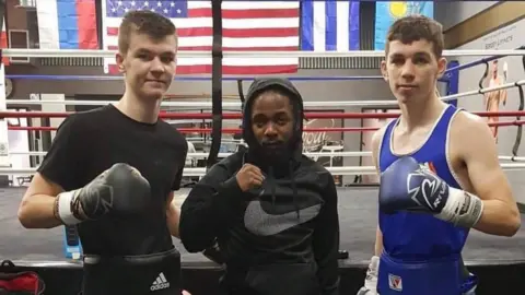 Team McKenna Aaron and Stephen McKenna stand with rapper Kendrick Lamar inside a Los Angeles boxing gym, with a boxing ring and national flags visible behind them.