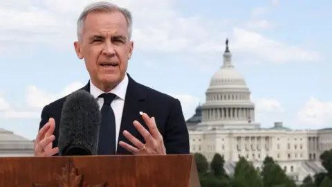 Canadian Prime Minister Mark Carney stands at a wood podium wearing a dark suit and white shirt. He is moving his hands and the US Capitol building is behind him. 