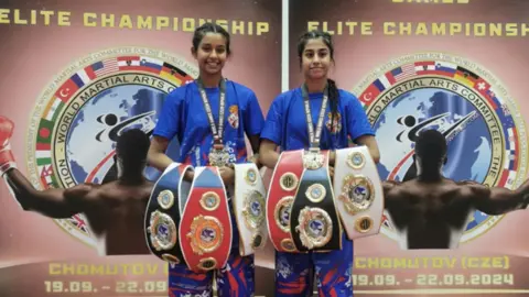 Family Dheep, 17 and Simran, 14 holding three belts each while standing in front of a kickboxing championship sign