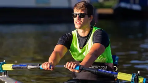 Nick B Images Xander Van der Poll rowing in a river, wearing a black shirt, green sports bib and black sunglasses