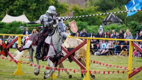 HES A knight with a patterned lance charges past another knight on his horse in a joust. There is a crowd watching in the background.