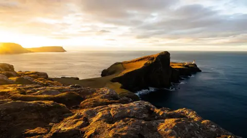 A dramatic coastal headland at sunset, with golden light hitting rocky cliffs that extend into the sea, and a lighthouse visible at the far end of the peninsula.