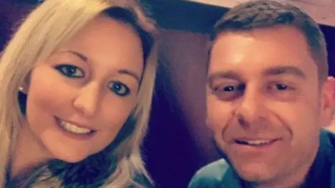 A selfie of Laura and Shane sitting close together indoors. Laura has light-colored hair and is wearing a coat with a furry collar. Shane is wearing a jacket over a dark top. Both are smiling at the camera.