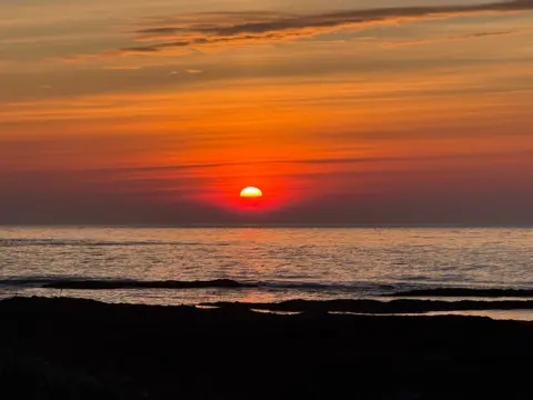 Suzanne Reid A vivid orange and red sunset over a calm sea, with the sun sitting just above the horizon and scattered clouds streaking the sky.