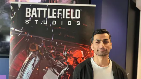 A man in a black cardigan over a white t-shirt stands in front of a "Battlefield Studios" poster. On it is a painterly image of a soldier inside a warplane with a cargo door open at the rear.