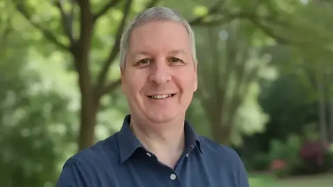 Jon Norman Garden Design A man with short grey hair is standing in a garden. He is wearing a navy blue polo shirt and is smiling at the camera. 