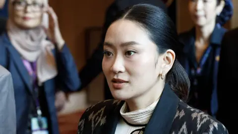 Close-up of Paetongtarn Shinawatra looking to the side. Her black hair is tied into a ponytail and she is wearing a patterned suit.
