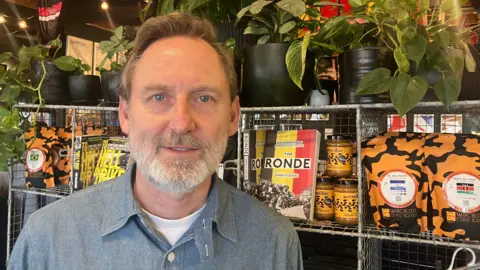 Image shows Neil Wyatt, a cafe owner in Winchester. He has light brown hair which is slicked over. He has a beard and is smiling at the camera. He is wearing a blue denim-look shirt with a white top underneath. Behind him is a metal shelf with little cubby holes. Each cubby hole is filled with a number of items the shop sells such as coffee beans, butter and books. On top of the cabinet is a number of green plants in black pots. 