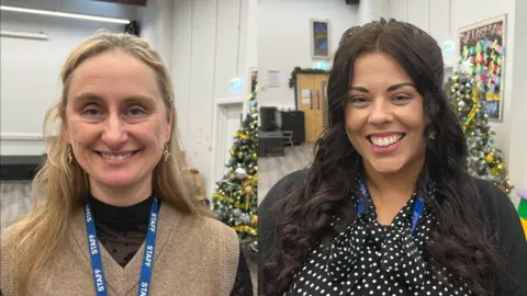 On the left-hand side is Hannah. She has blonde shoulder length hair and is smiling at the camera and wearing a blue lanyard. On the right is Stacey. She has long dark brown hair, and is also smiling in the camera. There are Christmas trees behind them.