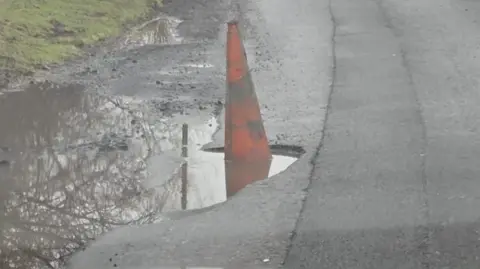 UGC A close-up of the large pothole on Margrove Road. It juts out from the side of the road, between the main carriageway and a grass verge. In this photo, it is flooded and a dirty orange traffic cone is partly submerged in the water.