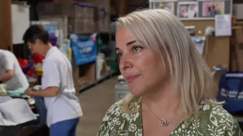 John Fairhall/BBC Svetlana Ryzhevska, in a green top and with white blonde hair, looking to the side of the camera in a warehouse.. Behind her volunteers are looking through donations. 