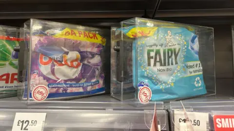 A mega pack of Bold liquid detergent pods in its own plastic container beside another XXL Fairy (non-bio) packet of liquid detergent pods also in a plastic container. There are only one of each item on the shelf and both priced at £12.50.