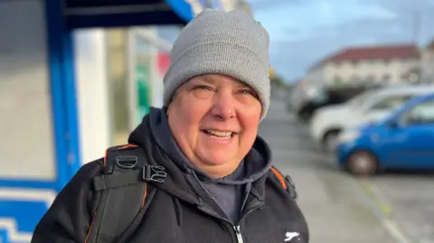 Nikki Ashman - A woman with a grey beanie hat on, wearing a black coat, with a back pack on her back. Under her coat is a blue hoodie. 