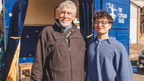 A young man stood next to his grandad in front of an old horsebox which has been converted into a stage 