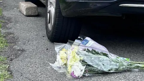BBC/Beth Parsons Two flower bouquets in plastic wrapping have been laid on a driveway in front of a car. The car's left back wheel can be seen in the picture.
