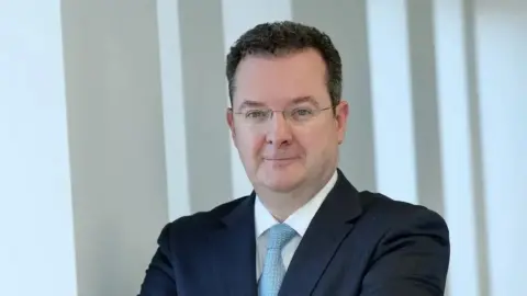 Kieran Donaghue has short dark hair and wire framed glasses. He is wearing a dark suit jacket, with a white shirt and blue tie