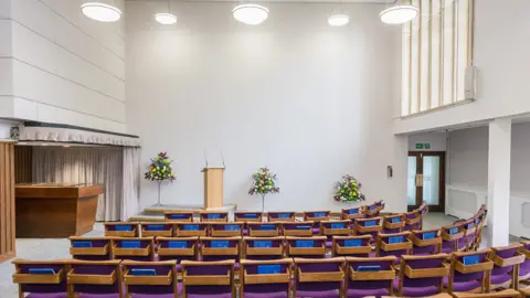 Worcester City Council The interior of Worcester Crematorium's chapel. Seats are arranged in front of a lectern and the room is lit by lights hanging from the ceiling.