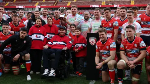 Getty A large group of Gloucester rugby players and mascots, along with Leicester players in the centre, holding a gold trophy up, stand around Ed Slater on a rugby pitch surrounded by empty stands post-match. 