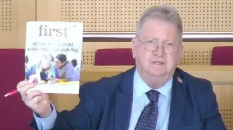 A man with glasses, a navy suit, light blue shirt. He is holding up a magazine or leaflet with children on the front of it.