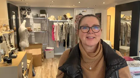 Caron Gardner, wearing glasses, with dark hair tied back, smiles at the camera as she stands in a clothing boutique shop.