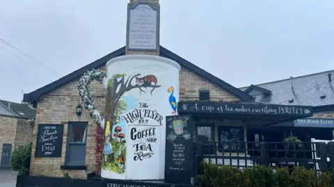 The side of a pub with a large white mug on the chimney breast. The building is brick with black tiled rooftops and signs advertising its tea and coffee shop. The mug is painted with scenes from Alice in Wonderland including Alice and a grinning cat in a tree