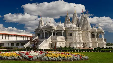 Getty Images An elaborate Hindu white stone temple with domes and turrets with gold on top. It is set against a blue sky with a colourful flowerbed in the foreground. 