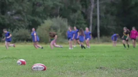 Young girls train at Nant Conwy Rugby Club. In the foreground are two rugby balls lying on the grass pitch. The players can be seen blurred in the background running and wearing blue bibs.
