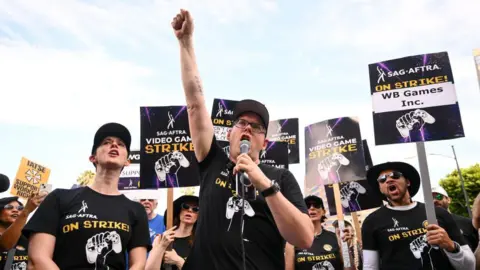 Getty Images A man stands with his fist raised as he speaks into a microphone. He's wearing a black t-shirt with a picture of a fist clenched around a gaming controller and the text 