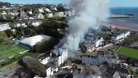 An aerial view of smoke billowing from the derelict hotel. The building is white and the roof is black with smoke.
