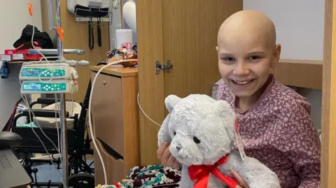 EACH Poppi Frisbi holding a large grey teddy bear while sitting on a hospice bed. She is smiling and wearing a pyjama top. To her right is hospital equipment.