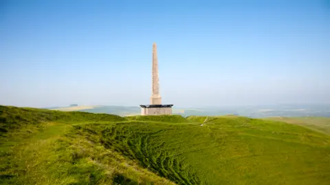 An obelisk with fences and netting at the base. The monument is surrounded by green fields. The image was taken on a sunny day.