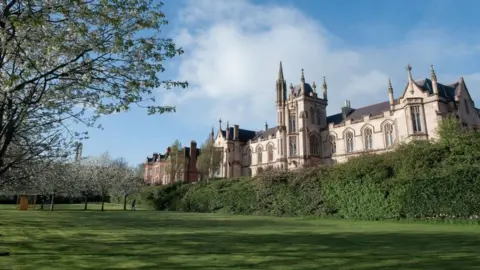 UU The university campus in Londonderry. A number of bushes and grassy areas can be seen in front of a Gothic-looking building with several large windows.