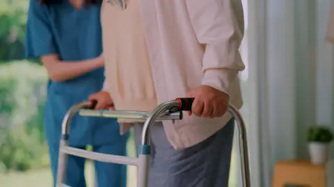 Image of a person using a silver walking frame while assisted by a person wearing a blue health worker's uniform