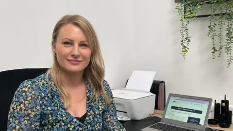 Sarah Jane sits at her desk with her laptop open. She has long blonde hair and is looking into the camera.