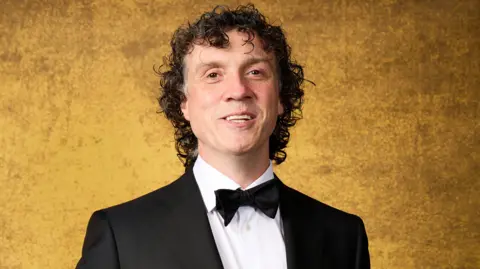 Getty Images Darragh Ennis is seen in a suit and bow tie in front of a gold background