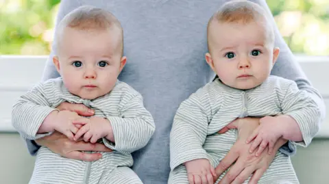 Two babies in green and white striped baby-grows. An adult is carrying them under either arm. 