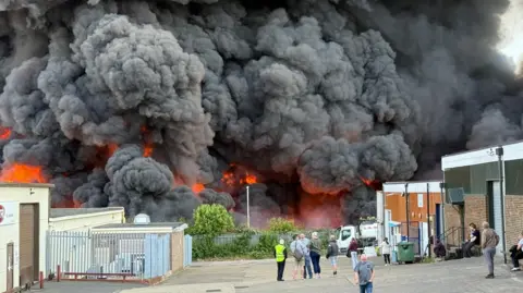 Steve Hubbard/BBC Large thick black smoke clouds with orange flame visible beneath. There is a boarded-up building to the right, and further small white buildings to the left. There are people standing in front of the fire, including one in a yellow hi-vis jacket.