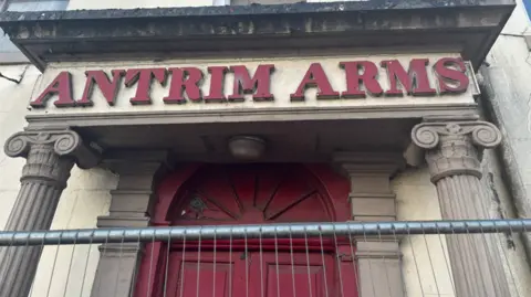 A large red sign reads Antrim Arms in front of a metal gate. 