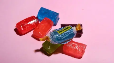 Seven individual Jolly Rancher Hard Candy sweets. All are unwrapped except a blue one. In total there are three red, two blue, one green and one purple sweet.