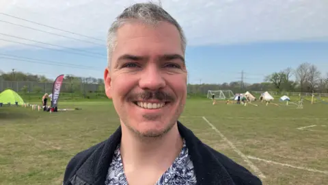 A man smiling with grey hair and a black moustache. He is wearing a floral shirt with a jacket over the top. In the background are several tents standing on a mown grass field.