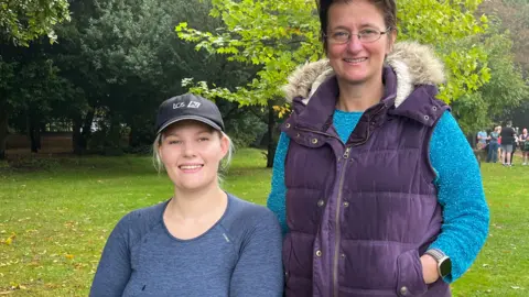 BBC/JULIA GREGORY Poppy May smiles into the camera. She is wearing a black baseball cap and blue long-sleeved top. Next to her in the grassy area stands her mum Helen Ghosh who has dark short hair and glasses and is wearing a turquoise jumper and a navy padded gilet with fur effect hood.