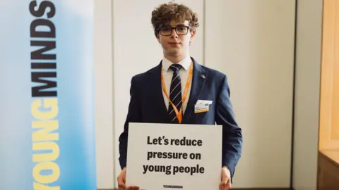 Stefano Di Rico A student in a blue suit and black pinstriped tie holding a sign that says "let's reduce pressure on young people". 