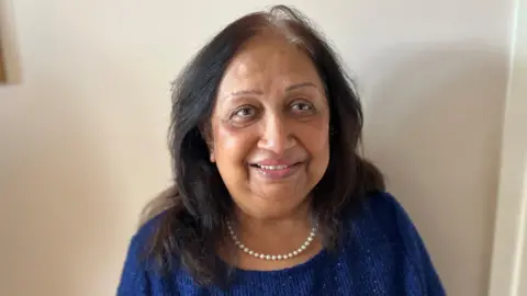 Ranjan Sodha, an older woman with black hair which is grey at the roots. She is smiling and wearing a royal blue knitted top with a pearl necklace.