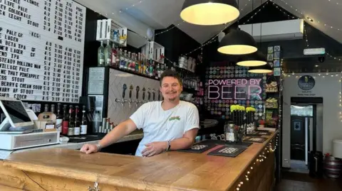 Julia Bryson/BBC A man in a white T-shirt with dark hair stands at a bar - there is a pink LED sign which reads 'powered by beer'.