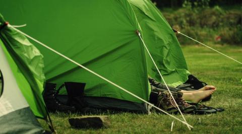 A generic image of a green tent in a grassy field with walking boots sat outside and a pair of feet poking out of the tent