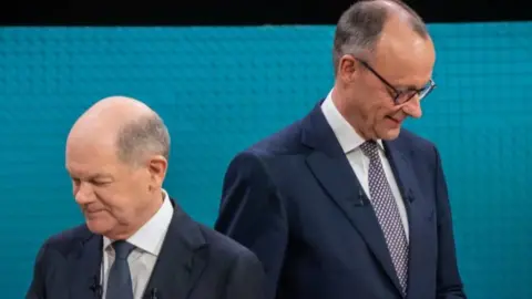 Friedrich Merz and rival Olaf Scholz standing next to each other. Both are wearing dark suits